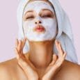 Beautiful young woman applying facial mask on her face. Skin care and treatment, spa, natural beauty and cosmetology concept, over pastel lilac background